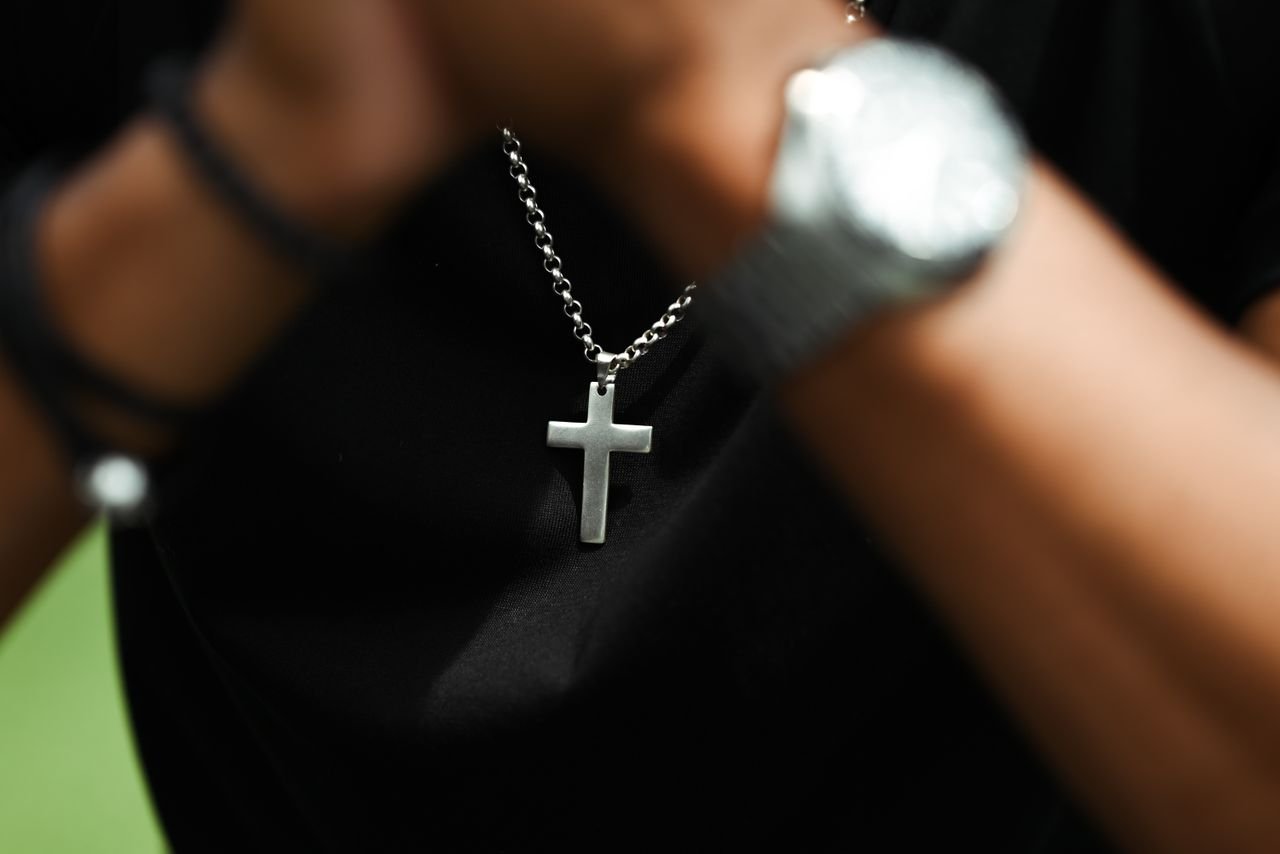 Men's Jewelry A close-up image of a silver cross pendant on a chain necklace worn by a person with a blurred watch in view.