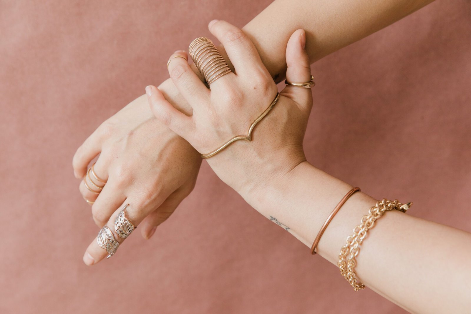 Elegant hands adorned with gold jewelry against a soft pink background, showcasing style and grace.